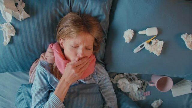 Beautiful 25s woman with scarf on neck coughs and blows her nose in napkin, woman covers herself with a warm blanket over her head. Flu and other respiratory diseases. Top view
