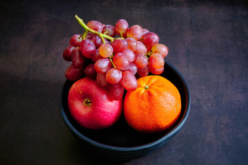 Red delicious fresh grapes, orange and apple on a black bowl. ready to eat. Juicy fruits background. Healthy eating, Fruit juice shop. 
