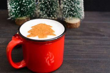 Christmas red cup of coffee with fir tree pattern. New Year beverage over dark wooden background with copy space. Mag of cuppuccino with art in shape of Christmas tree and decorations. Selective focus