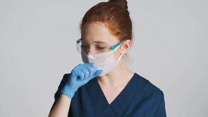 Young doctor in protective eyeglasses, mask and medical gloves coughing over white background