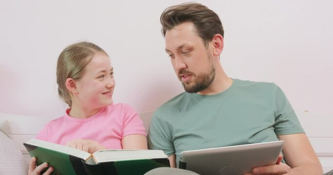 Close up of father, holding a tablet, and laughing teen daughter sitting on the sofa, and looking at the photo album.