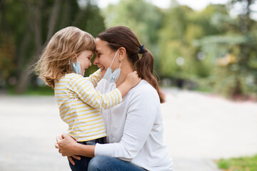 Mom and child girl with medical face lowered mask hugging outdoors