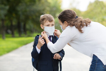 Mother wearing on medical mask puts a safety mask on her schoolkid son's  face outdoors. schoolboy wearing mask with backpack going to school.