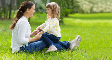 happy Mom and her child daughter sitting in the grass and having fun togetherss in sunny day outdoors. copy space
