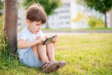 Cheerful child sitting on the grass looks cartoons in the phone in the summer at sunset. Cute boy having fun in nature