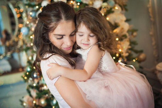 A beautiful young mother holds her little daughter in her arms and they hug each other tightly. Their eyes are closed from tenderness. Christmas picture, tree and lights in the background.