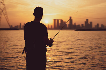 During the enchanting sunset, a young angler stands on the beach, casting their line while gazing...