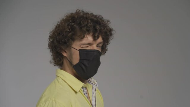 Curly man in a medical mask winks at the camera and raises his eyebrows. Playful look in a protective mask. Flirting on a quarantine concept. High quality 4k footage.
