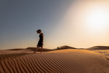 a young woman gracefully traversing the desert terrain, her footsteps etched into the soft, golden sands of a towering dune. - obrazy, fototapety, plakaty