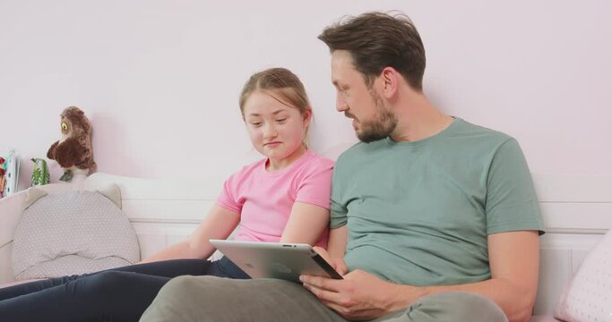 Father and teen daughter sitting on the sofa in living room, browsing the online store, enjoying weekend. Father asks what she thinks about some of articles.
