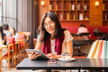 Advertising, Business, Technology Concept - Beautiful young asian woman holding digital tablet and looking at camera with smile while sitting in a modern cafe and near the girl is colorful bags.