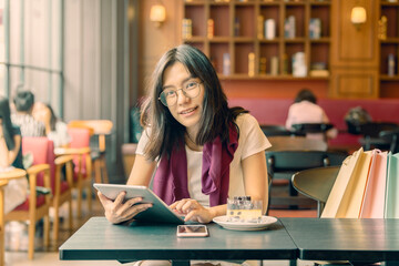 Beautiful young asian woman holding digital tablet and looking at camera with smile while sitting in a modern cafe and near the girl is colorful bags and background look old or vintage style...