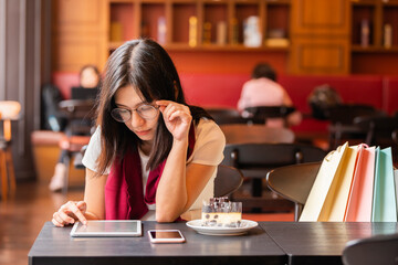 Lifestyle, Technology Concept - Asian woman doing online shopping through tablet computer and near the girl is mobile phone, cake, colorful bags in a modern cafe.