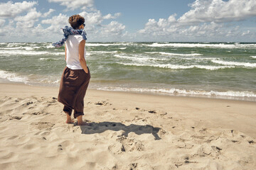 Middle aged woman standing on the beach looking at waving sea on windy summer day