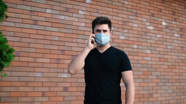 Portrait of a man dressed in black, talking on the phone. Young man with mask on his face talking on the phone. In the background is a brown brick wall. - Powered by Adobe