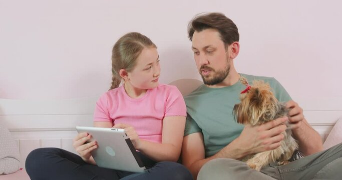 Father, with a dog on his palm, and teen daughter, holding a tablet, are sitting on the sofa in living room, browsing the online store, enjoying weekend. Father agrees to choose the thing, his