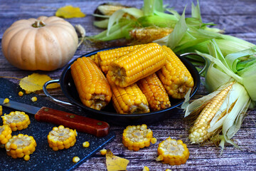 Autumn composition of boiled corn on a plate, ears of corn and pumpkin on a dark wooden table. Seasonal harvest