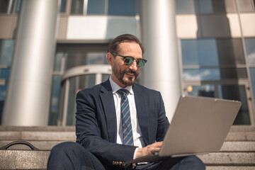 Successful businessman works on laptop sitting on steps of business center. Smiling man wearing business suit and sunglasses. High quality photo.