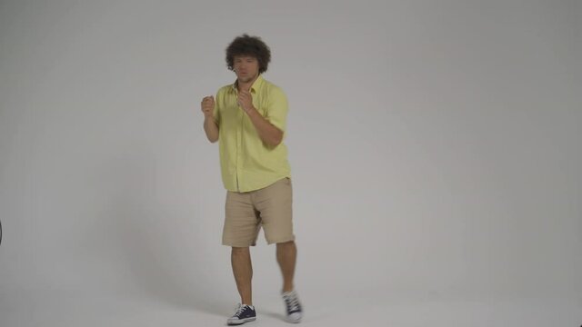 Young man happily and victoriously dances in the studio. Curly joyful guy on an isolated background. High quality 4k footage.