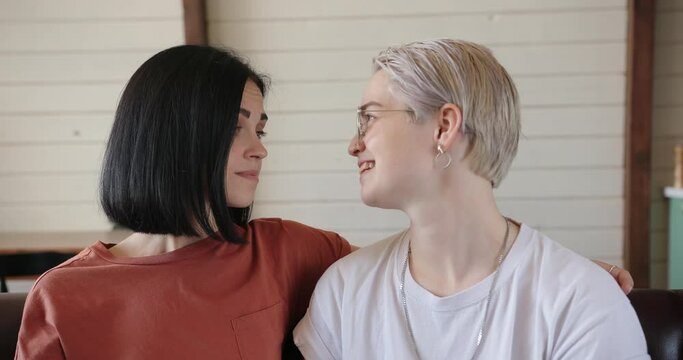 Tattooed lesbians in brown and white t-shirts kiss and hug sitting on leather sofa at home with white wall slow motion closeup