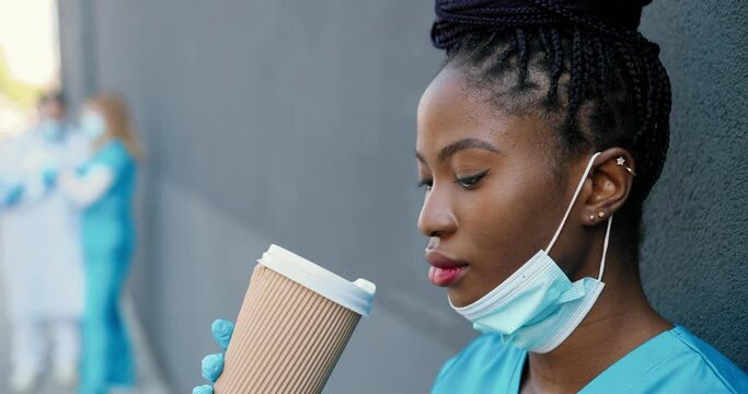 Close up tired young African American woman doctor taking off medical mask and sipping hot drink while resting and leaning on wall outdoor. Pretty female nurse drinking coffee and rest after hard work