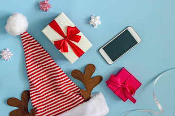 Christmas greetings online flat lay. Top view on a blue background Santa hat in red and white stripes, smartphone, gift boxes with bows, copy space. Phone congratulations merry christmas background