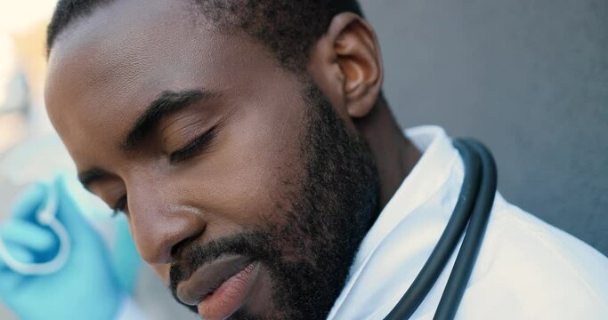 Tired Young Sad African American Man Doctor Taking Off Medical Mask And Resting While Leaning On Wall. Male Medic Rest After Hard Work. Lost Life. Difficult Day Of Dissapointed Physician. Close Up.