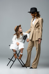 elegant mother and daughter in white and beige outfits and hats posing on chair and looking at each other on grey background