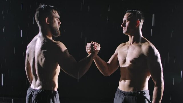 Two focused topless men shake hands amidst the raindrops and splashing water. Picture taken in the studio on a black background. Slow motion. Close up.