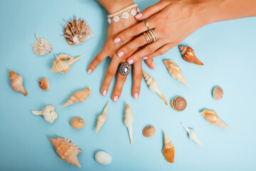 beautiful woman hands with pink manicure holding sea shells, lot of rings on fingers on blue background, luxury jewelry concept