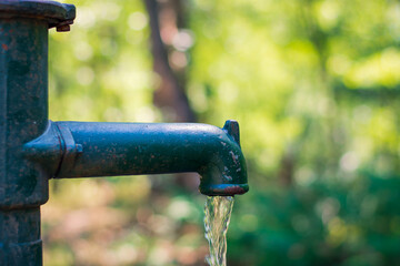 Manual old green and rusty water lever pump with water pouring out of the spout with a blurred green background