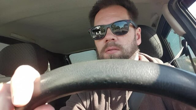 bearded man with glasses driving a car, taking a selfie with a smartphone camera