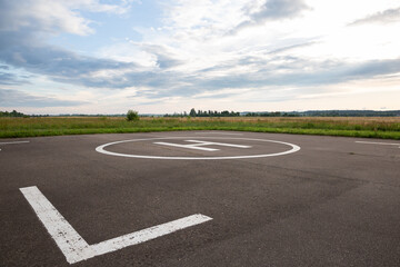 Fototapeta premium A closeup of an asphalt-covered helipad with a special symbol in the center for helicopter landing, against the backdrop of a green field and a cloudy evening sky.