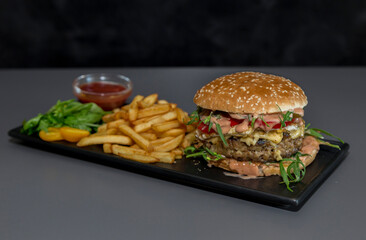 One big tall classic hamburger burger cheeseburger with french fries on black stone plate. Classic burger, french fries, sauce and greens on a black plate in a restaurant.