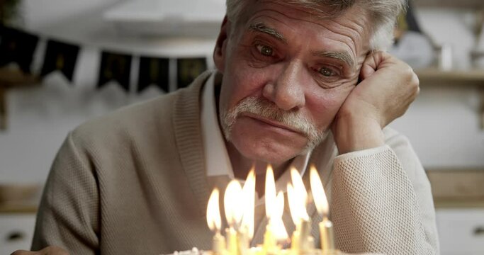 Close-up of deeply disappointed senior man's face sitting alone at kitchen table in front of cake with lit candles, celebrating birthday without relatives, suffering from loneliness, thinking of life.