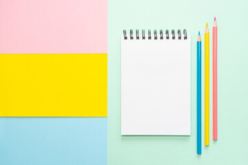 Spiral notebook with blank white sheet and colored pencils on multi-colored paper. Concept of office, school, creativity, learning. Flat lay, copy space, minimalism. Pink, blue and yellow shades.