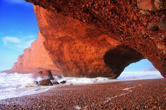 Legzira beach, Morocco, Northwest Africa. Beautiful nature view, famous red mountain arch in the light of the setting sun, stormy Atlantic ocean, pebble coast, sea surf and blue sky.
