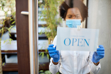 Black waitress with protective face mask and gloves hanging open sign at entrance door.