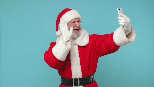 Glad Santa Claus man in Christmas hat red suit coat white gloves glasses doing selfie on mobile phone gadget isolated on blue color background studio. Happy New Year celebration merry holiday concept
