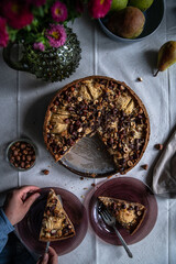 Fototapeta na wymiar Top view of frangipani tart with pears, hazelnuts and chocolate, plates, fresh pears and vase with flowers on linen tablecloth .