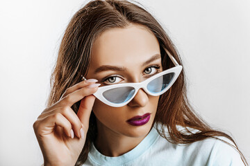 Beautiful young brunette girl with green eyes with fashion makeup with purple lips on an isolated gray background. The woman in blue sunglasses seriously looking at the camera. Beauty concept.