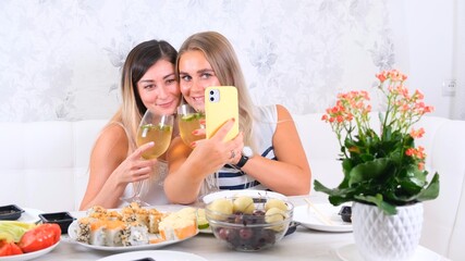 Obraz na płótnie Canvas friendship and party concept - two young beautiful girlfriends drink wine and take selfie on smartphone in a cozy kitchen at the table, selective focus
