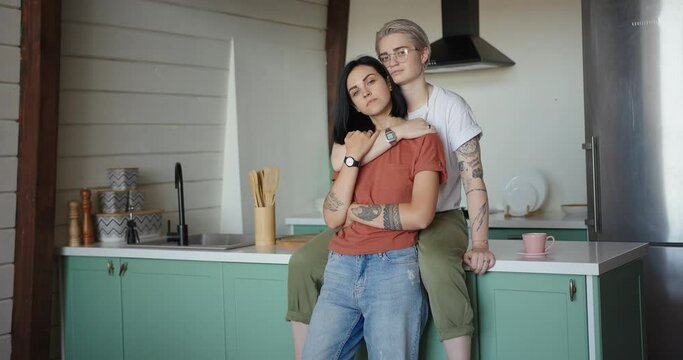 Tattooed lesbian with fair hair and glasses hugs brunette sitting on kitchen table near sink and posing at home slow motion