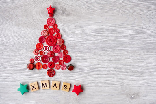 top view, on a white wooden background, a Christmas tree is laid out with red buttons, and the Xmas inscription is made of wooden letters at the bottom. Space for text on the right.