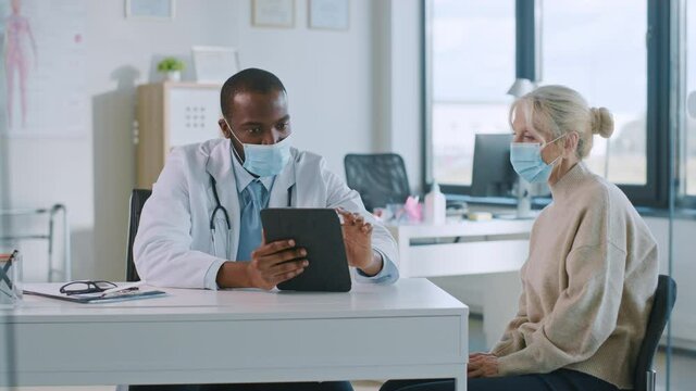 African American Doctor in Protective Mask is Reading Medical History of Senior Female Patient During Consultation in a Health Clinic. Physician Using Tablet Computer in Hospital Office.