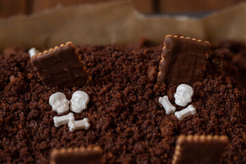 Kuchen als Friedhof mit Gräbern und Skeletten aus Schokokrümel, Creme und Banane auf Tisch aus Holz und dahinter Spinnweben