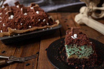 Kuchen als Friedhof mit Gräbern und Skeletten aus Schokokrümel, Creme und Banane auf Tisch aus Holz und dahinter Spinnweben
