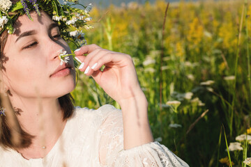 A beautiful woman in a white dress in the boho style with a wreath on her head sitting in a field in flowers. The concept of beauty, free life and naturalness