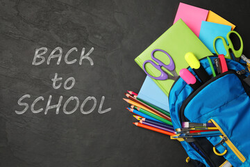 Text Back To School and backpack with different stationery on black chalkboard, top view