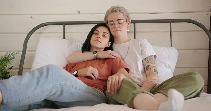 Tattooed dressed lesbian couple in love lies on bed with metal back and white pillows and hugs against wooden wall slow motion